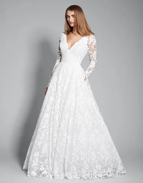 A woman modeling a long-sleeve white lace wedding gown with a deep V-neckline and a full A-line skirt, photographed against a neutral gray studio background.