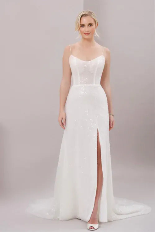 A woman modeling a white sleeveless wedding gown with thin straps, a fitted lace bodice, a high front slit, and a slight train against a plain light gray studio background.