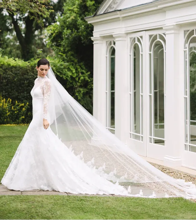 Woman in a white wedding dress with a long veil standing in front of a white building with arched windows.