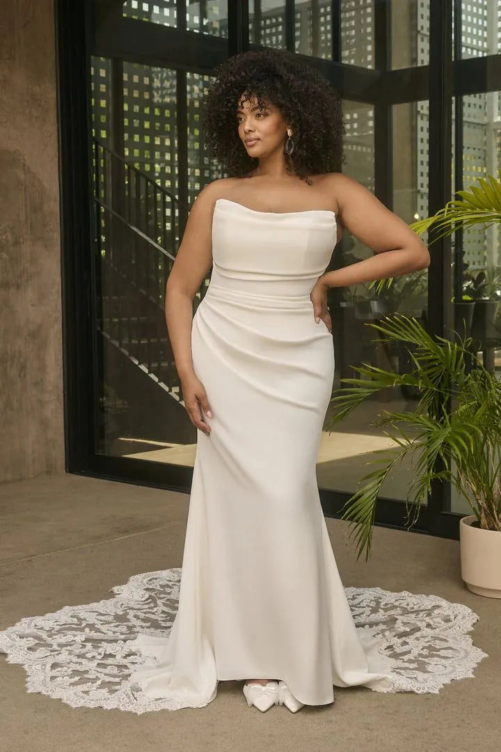 A woman modeling a strapless white mermaid-style evening gown with a fitted bodice, draped waist, and flared skirt, standing indoors near large black-framed windows and a potted plant.