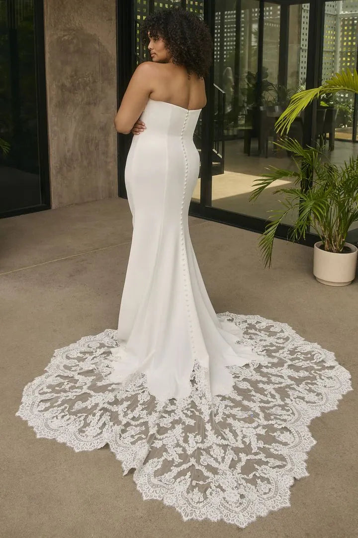 Woman wearing a strapless white wedding dress with a lace train in an indoor setting.