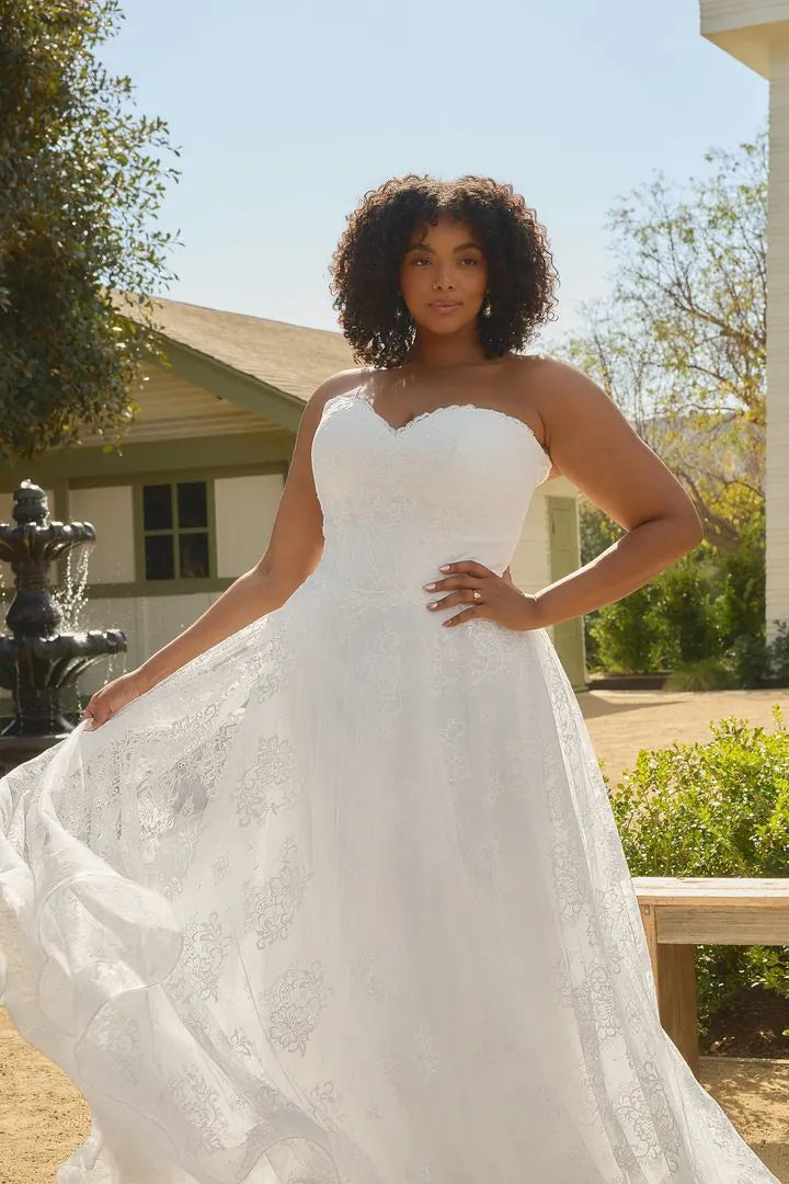 A woman wearing a strapless white lace wedding gown with a sweetheart neckline and full skirt, standing outdoors in a sunny garden setting.
