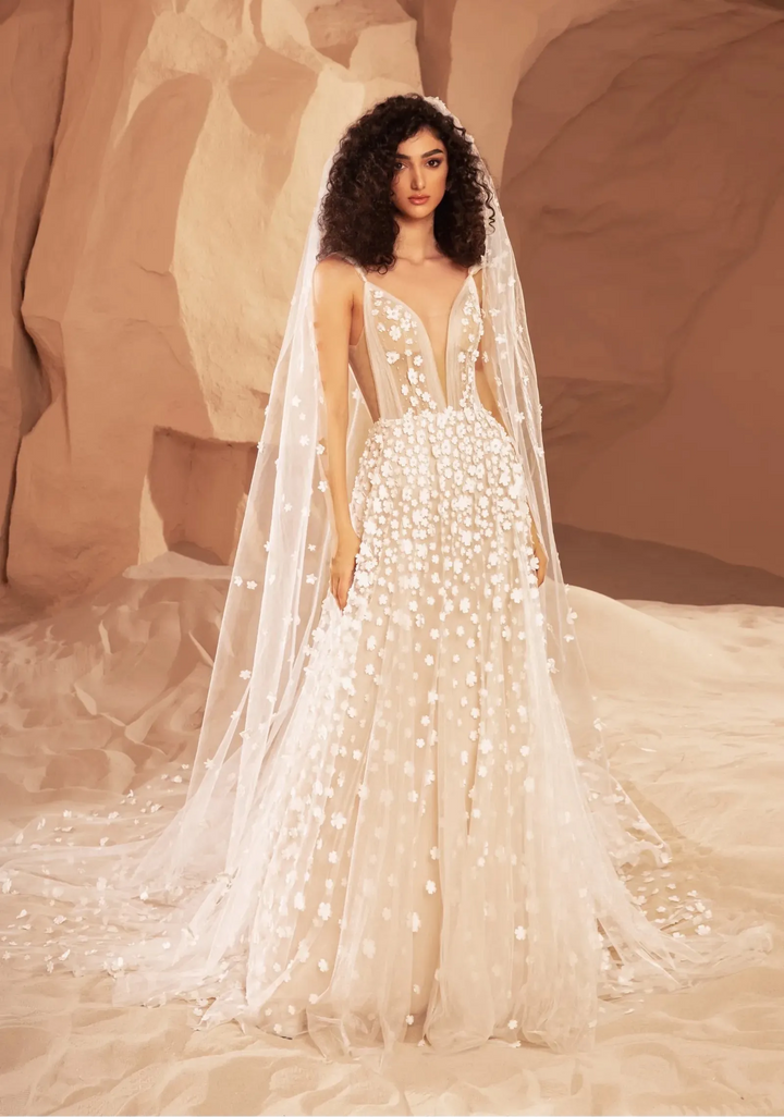 A model wearing a white beaded wedding gown with a long sheer veil, posed in a desert canyon setting with warm beige rock formations.