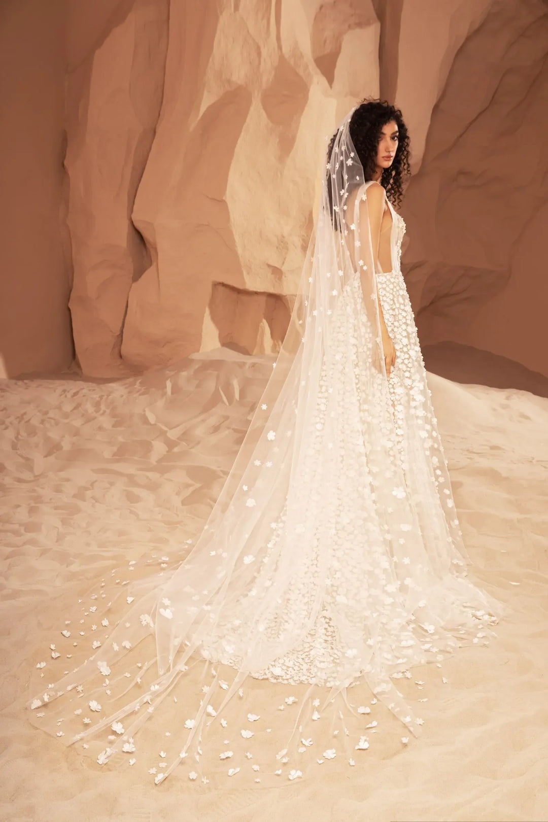 A woman wearing a long white bridal gown with a dramatic train and veil, posed in a warm desert canyon setting with beige rock formations.