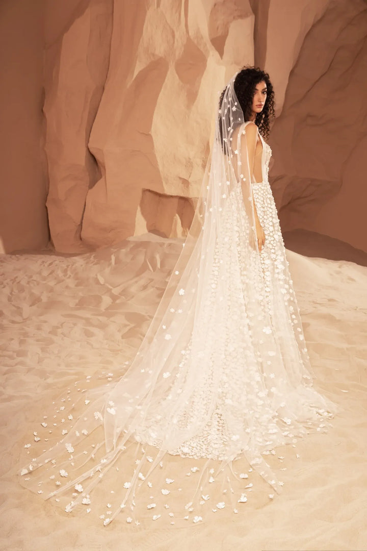 A woman wearing a long white bridal gown with a dramatic train and veil, posed in a warm desert canyon setting with beige rock formations.