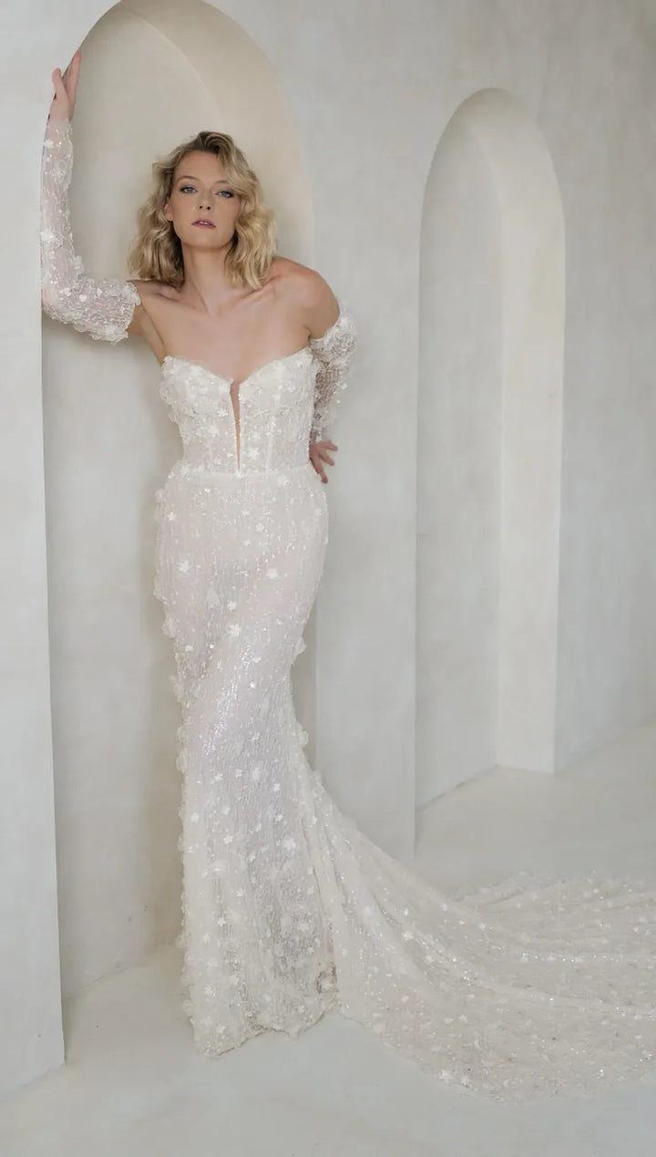 A woman modeling an off-the-shoulder, strapless mermaid-style wedding gown covered in sparkling beadwork and sequins, posed in a softly lit architectural setting with arched alcoves.