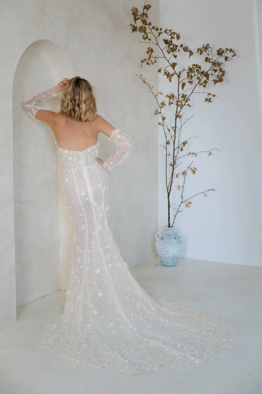 A woman stands with her back to the camera wearing a fitted off-the-shoulder wedding gown with long sheer sleeves and a long train, posed in a bright white interior with a decorative vase of dried branches.