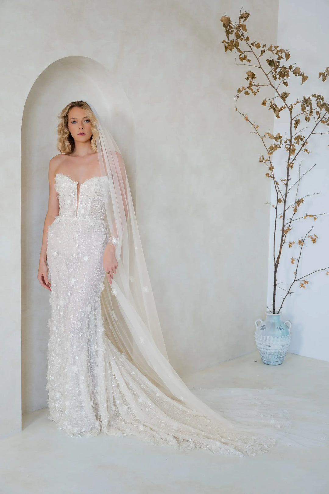 A woman wearing a strapless ivory lace wedding gown with a long sheer veil stands in a softly lit room with a curved wall niche and a vase of dried branches beside her.