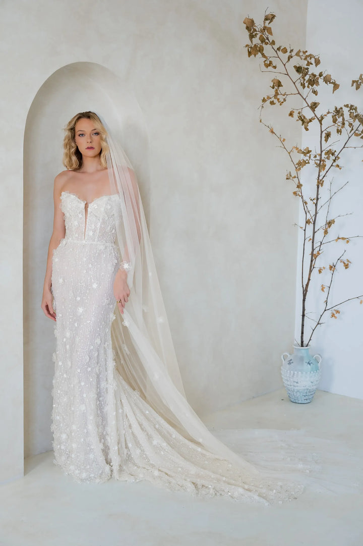 A woman wearing a strapless ivory lace wedding gown with a long sheer veil stands in a softly lit room with a curved wall niche and a vase of dried branches beside her.