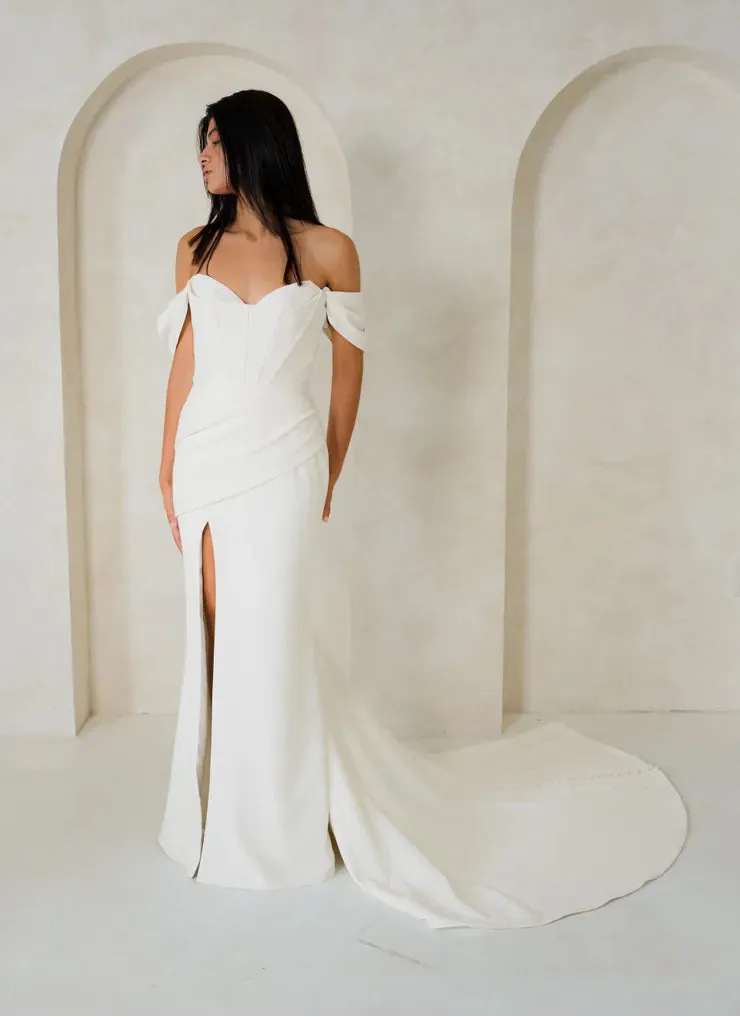 A woman wearing an off-the-shoulder floor-length wedding gown with a thigh-high slit and long train, standing in a minimalist cream-colored room with arched wall details.