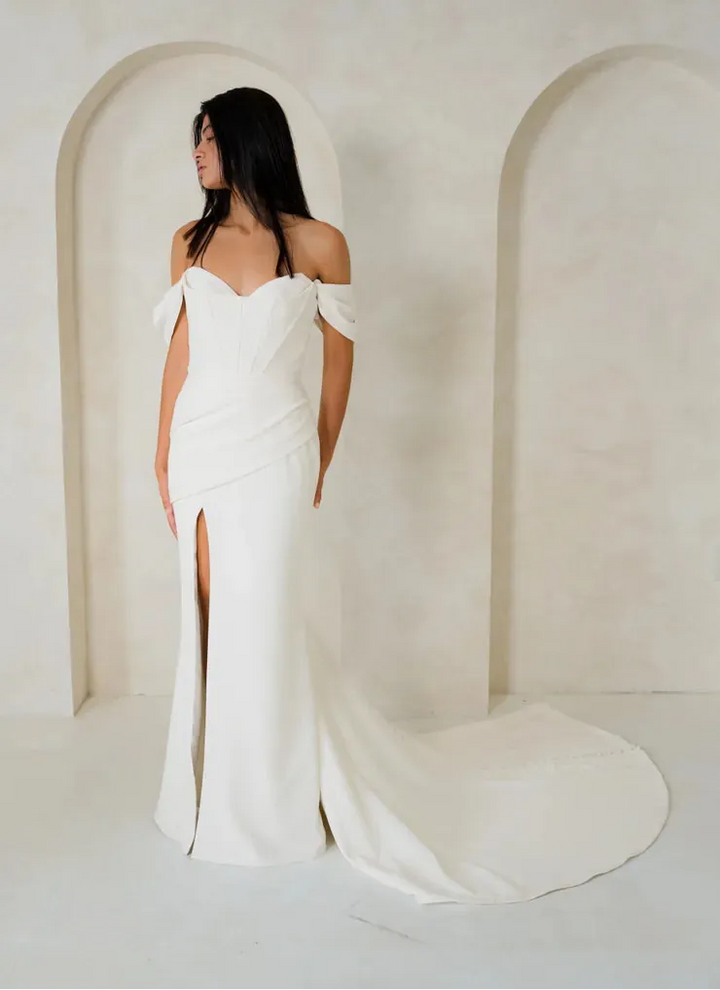 A woman wearing an off-the-shoulder floor-length wedding gown with a thigh-high slit and long train, standing in a minimalist cream-colored room with arched wall details.