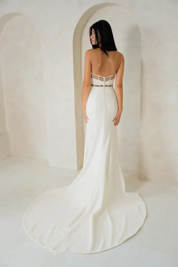 A woman stands with her back to the camera wearing a fitted white wedding gown with a low open back, strappy detailing at the waist, and a long train in a softly lit cream-colored architectural setting with arches.