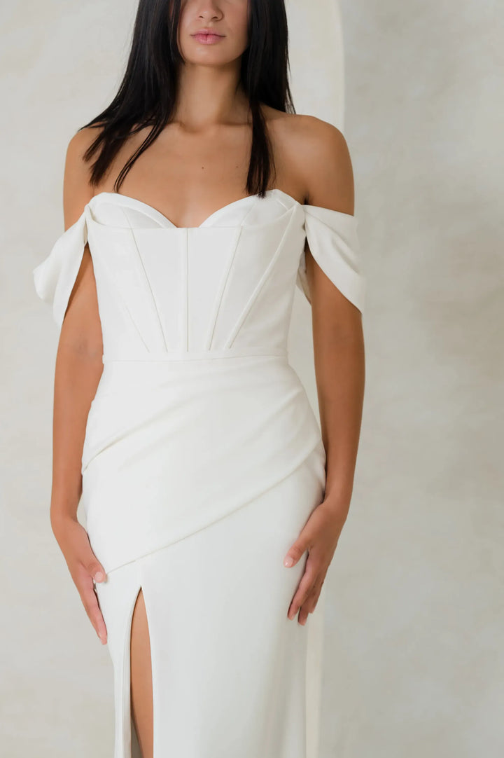 An off-the-shoulder white gown with a sweetheart neckline, structured corset-style bodice, and a high thigh slit, shown on a model against a neutral studio backdrop.