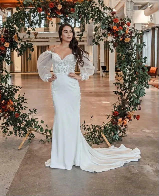 A woman stands in a large industrial loft wearing a fitted white wedding gown with sheer balloon sleeves and a long train, posed in front of a floral arch made of greenery and orange flowers.
