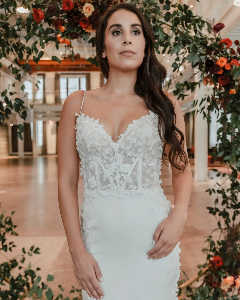 A woman wearing a white lace wedding dress with thin straps and a fitted bodice, standing in a decorated indoor venue with floral arches.