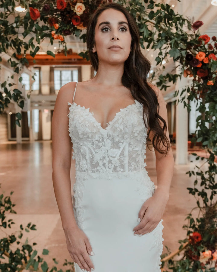 A woman wearing a white lace wedding dress with thin straps and a fitted bodice, standing in a decorated indoor venue with floral arches.