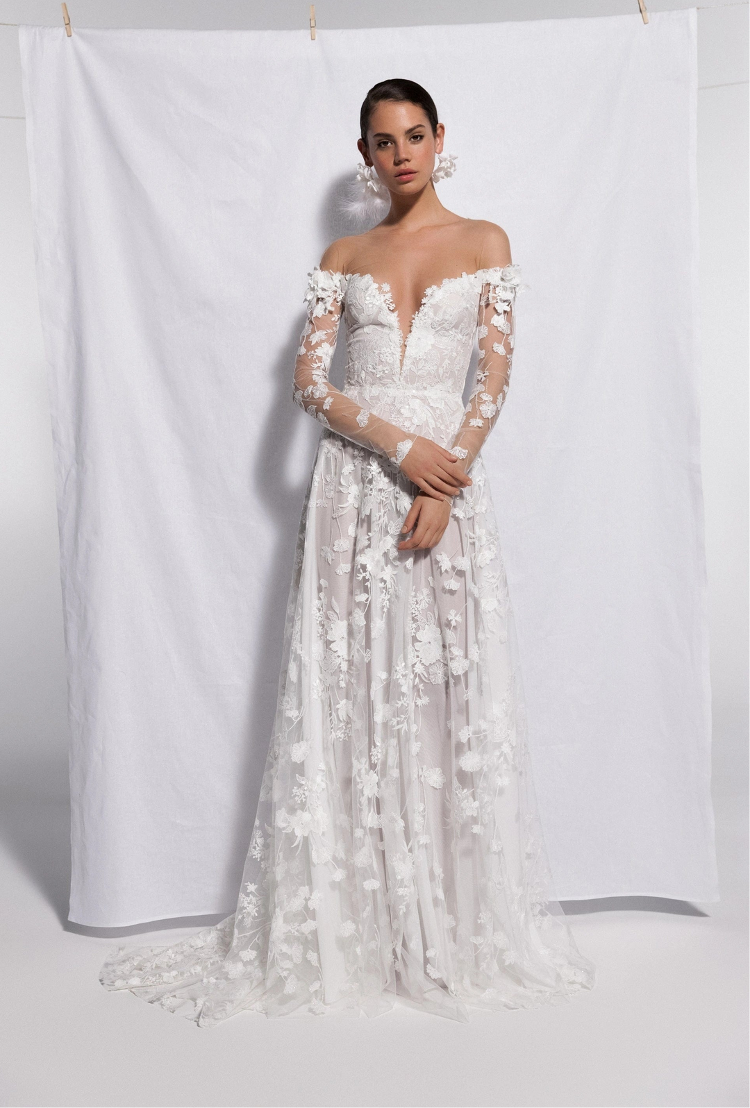 A woman models an off-the-shoulder white lace wedding gown with sheer long sleeves and a full skirt, standing in front of a white fabric backdrop.