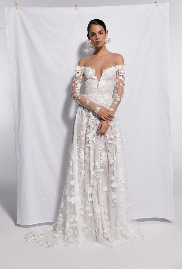 A woman models an off-the-shoulder white lace wedding gown with sheer long sleeves and a full skirt, standing in front of a white fabric backdrop.