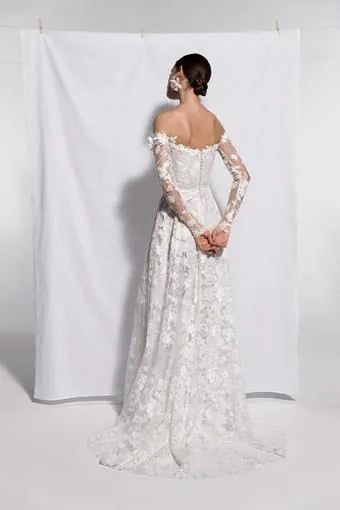 A woman modeling a white off-the-shoulder lace wedding gown from the back, standing in front of a white fabric backdrop.