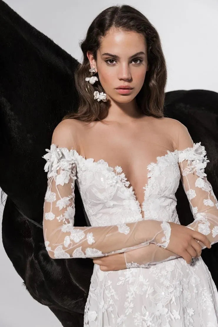 A woman modeling an off-the-shoulder white lace wedding dress with sheer long sleeves and floral applique, posed against a black feather backdrop.