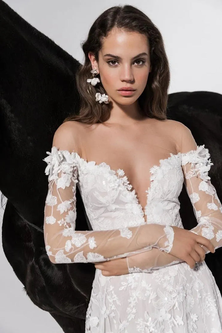 A woman modeling an off-the-shoulder white lace wedding dress with sheer long sleeves and floral applique, posed against a black feather backdrop.