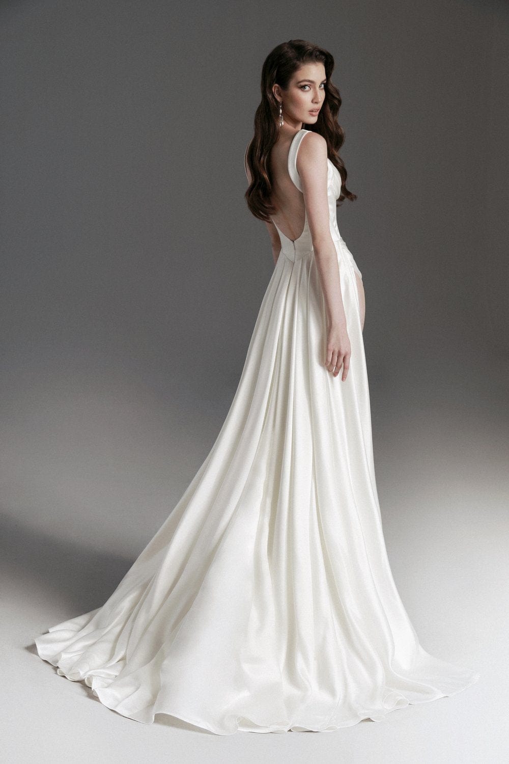A woman modeling a floor-length white wedding gown from the back, showing a deep V back, thin straps, and a flowing satin train against a dark gray studio background.