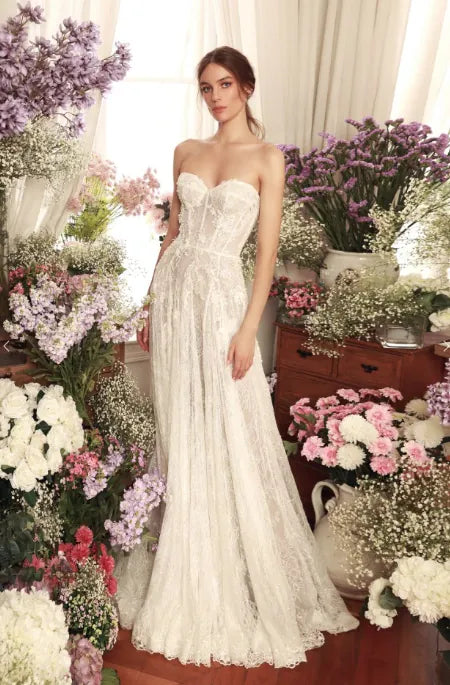 A woman wearing a strapless lace wedding gown stands indoors among abundant floral arrangements in white, pink, and purple tones. The dress has a fitted lace bodice and a full flowing skirt with a train.