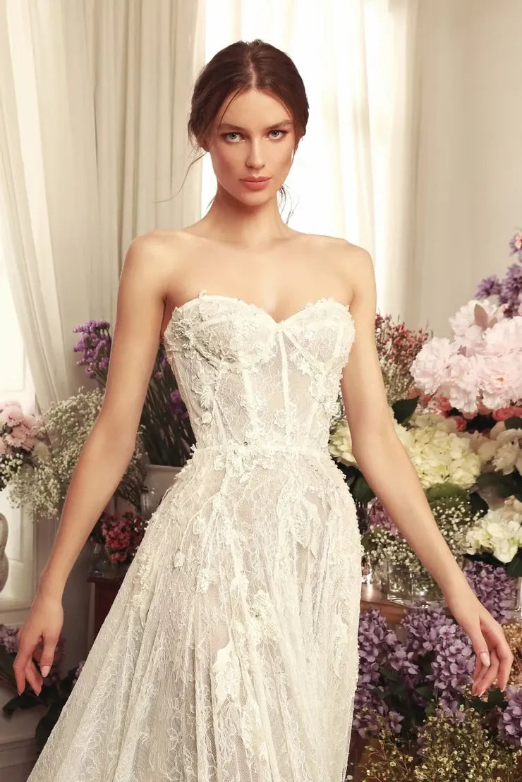 A woman modeling a strapless white lace wedding gown with a fitted corset-style bodice and full skirt, posed in front of cream curtains and floral arrangements.