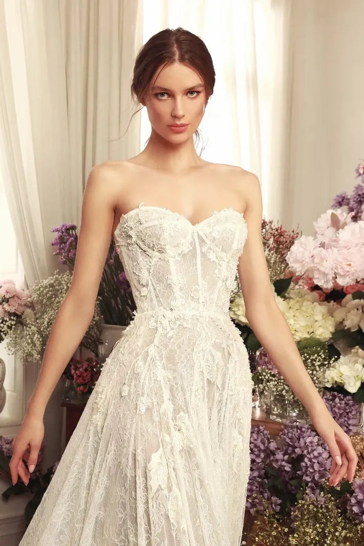 A woman modeling a strapless white lace wedding gown with a fitted corset-style bodice and full skirt, posed in front of cream curtains and floral arrangements.