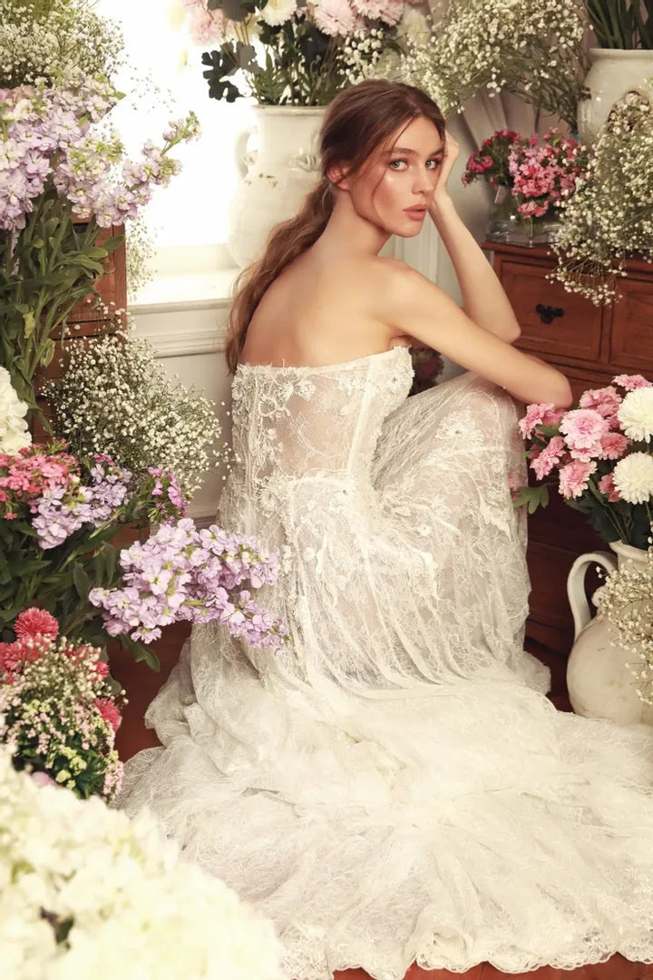A woman wearing a strapless white lace wedding gown sits in a floral room surrounded by pink, purple, and white flowers.
