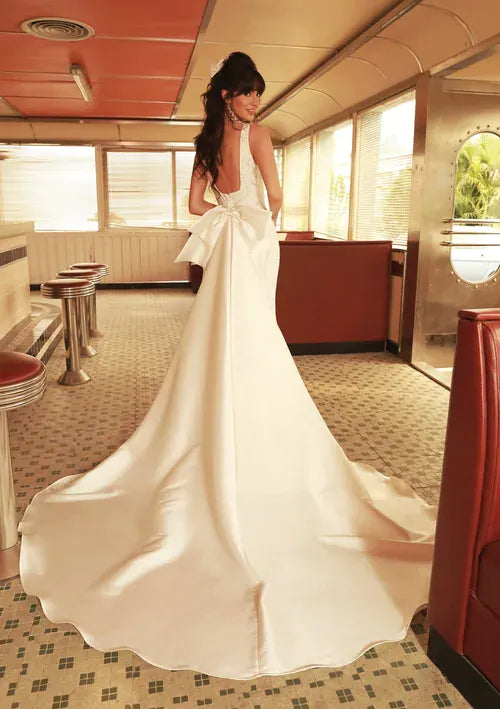 A woman wearing a white satin bridal gown with a dramatic train, photographed from behind inside a retro-style diner or train car interior.