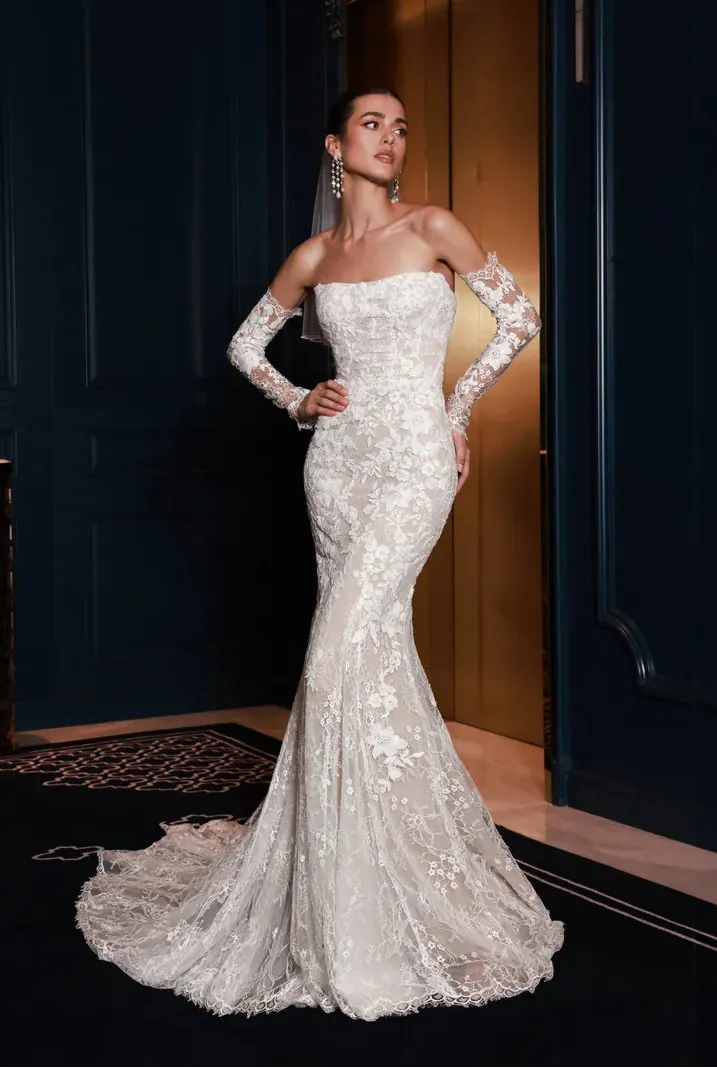 A woman modeling a strapless off-the-shoulder lace mermaid wedding gown with long sheer sleeves and a train, posed in front of dark navy paneled doors and a gold-toned interior.
