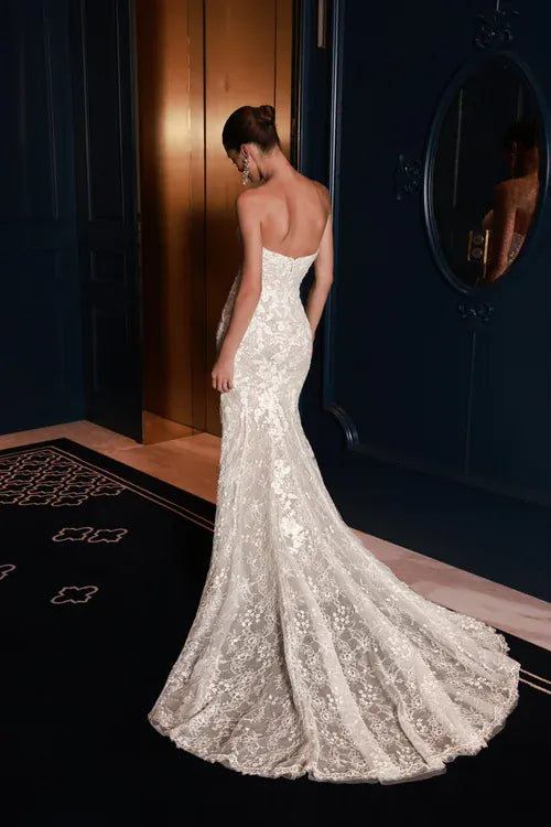 A woman wearing a strapless lace mermaid-style wedding gown is shown from the back in an elegant indoor setting with dark navy walls, a mirror, and a black patterned rug.