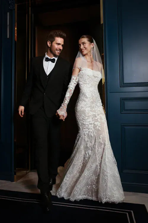 A bride in a fitted white lace mermaid wedding gown with long sheer sleeves and a matching veil walks hand-in-hand with a groom in a black tuxedo and bow tie in front of dark blue paneled doors.