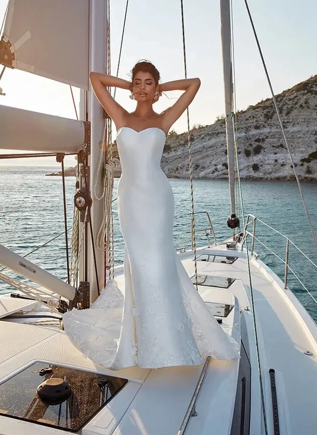 A woman wearing a strapless white wedding gown stands on the deck of a sailboat with blue water and a rocky coastline in the background.