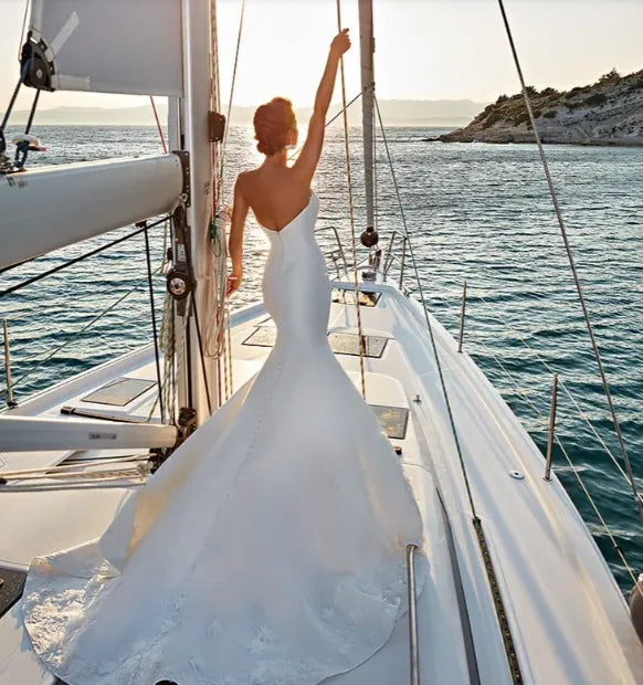 A woman wearing a white sleeveless mermaid-style wedding gown stands on the deck of a sailboat, with her back to the camera and one arm raised toward the rigging. The boat is on open water with a rocky shoreline in the distance under warm sunlight.