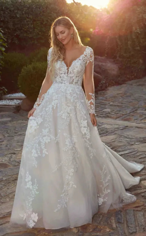 A woman wearing a long-sleeve A-line wedding dress with floral lace applique and a deep V-neckline, standing outdoors on a stone path with warm sunset light.