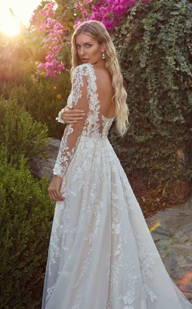 A woman wearing a long-sleeve lace wedding dress with an open back, standing outdoors in front of greenery and pink flowers at sunset.