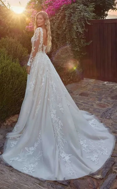 A woman wearing a long-sleeve white lace wedding gown with a full train, photographed outdoors on a stone path with warm sunset light and garden foliage in the background.