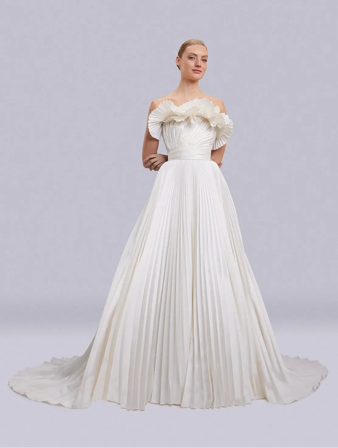 An elegant off-the-shoulder A-line wedding gown in ivory, featuring ruffled detailing on the bodice and a full pleated skirt with a long train, modeled against a light gray studio background.
