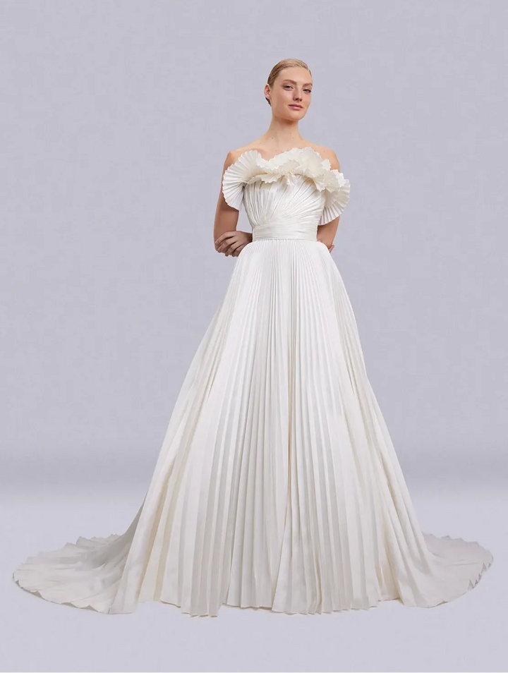 An elegant off-the-shoulder A-line wedding gown in ivory, featuring ruffled detailing on the bodice and a full pleated skirt with a long train, modeled against a light gray studio background.