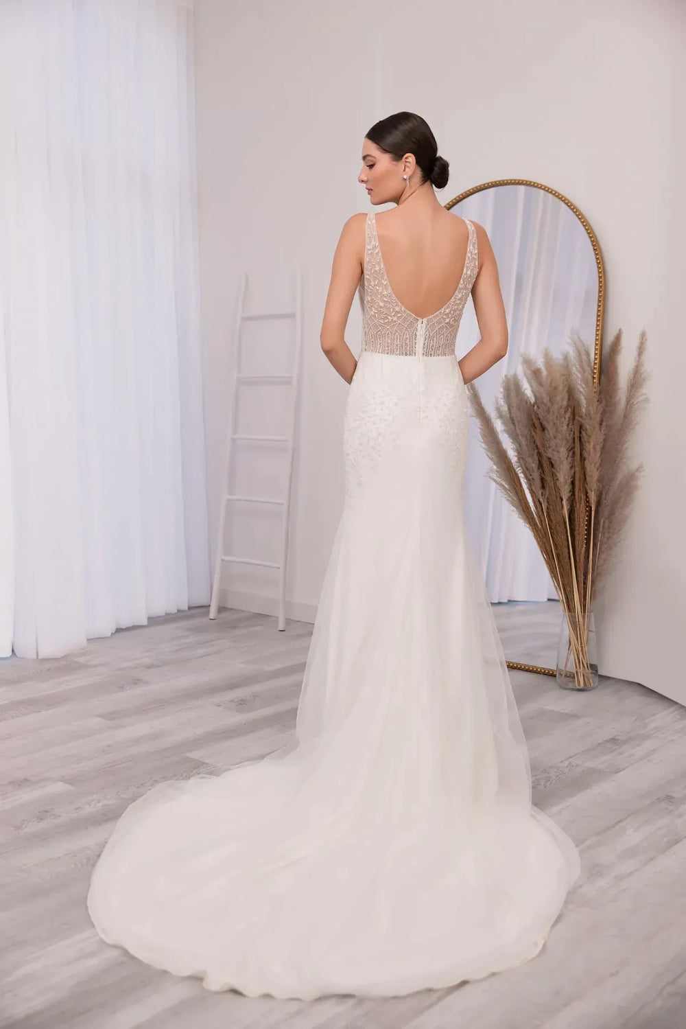 Woman in a white wedding dress with an open back standing in a room with a mirror and decorative plants.