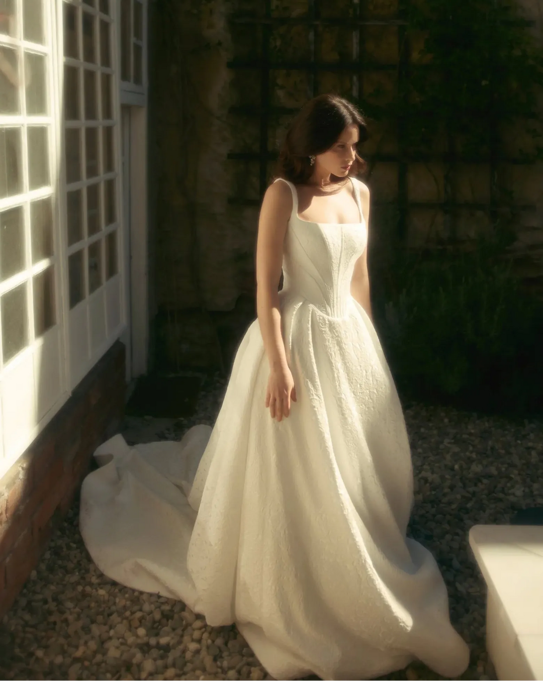 Woman in a white dress standing in a sunlit outdoor setting with plants and a building.