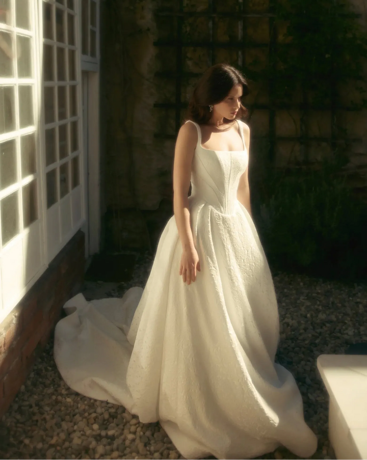 Woman in a white dress standing in a sunlit outdoor setting with plants and a building.