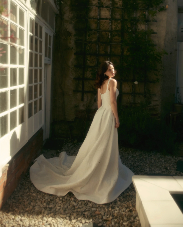 Woman in a white dress standing in an outdoor setting with greenery and a building.