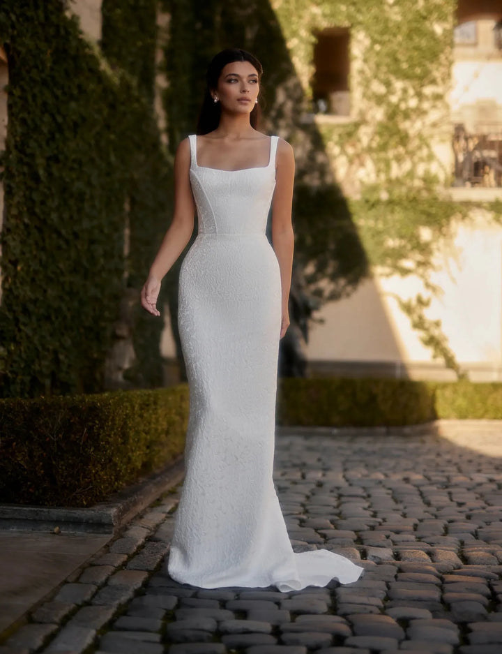 Woman in a white wedding dress standing on a cobblestone path with greenery and architecture in the background