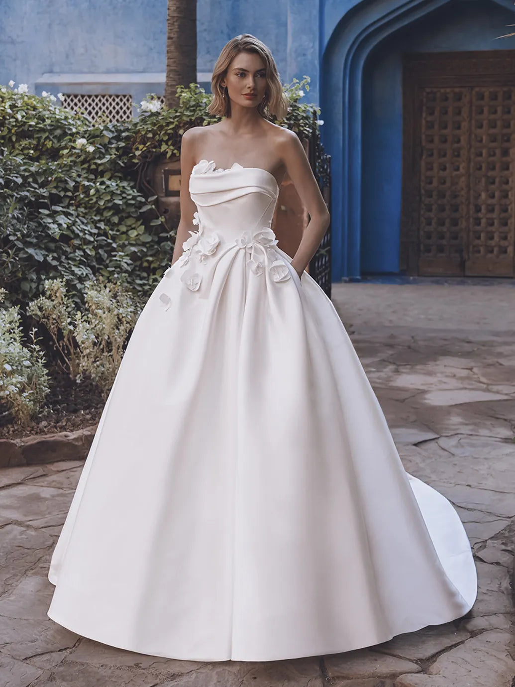 Woman in a white strapless gown standing outdoors with a blue architectural structure in the background.
