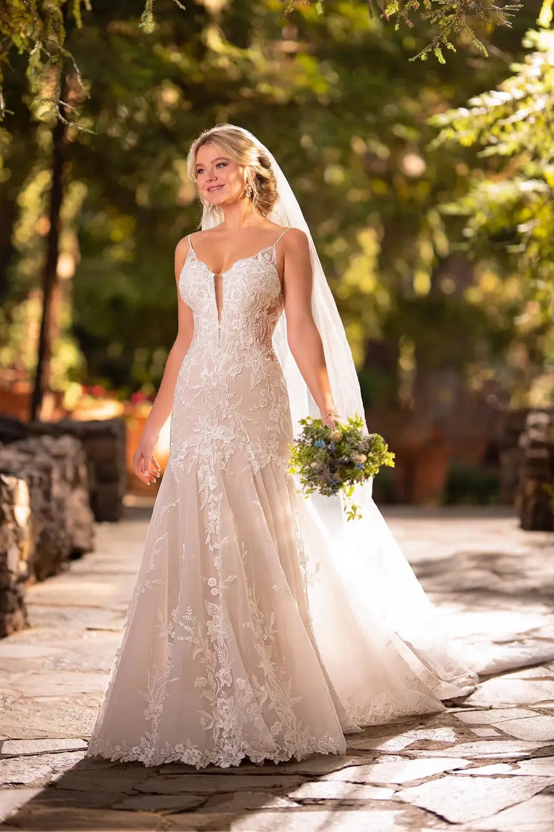 A bride wearing a strapless lace wedding gown with a deep V-neckline and a long sheer veil, holding a small bouquet outdoors on a stone path with trees in the background.