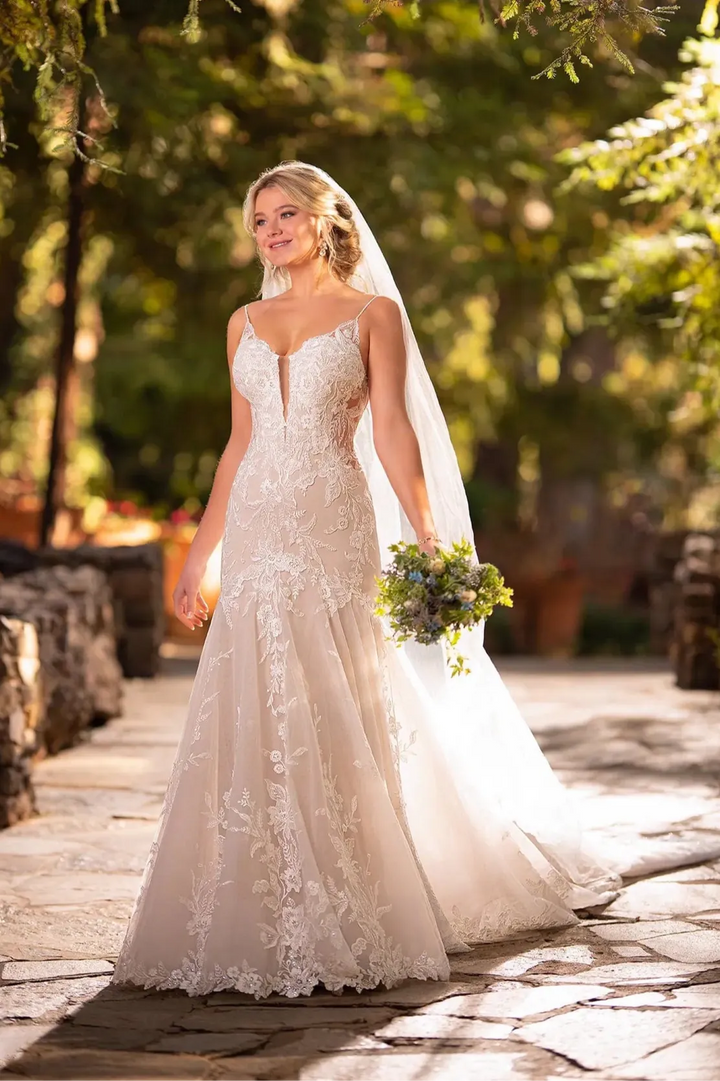 A bride wearing a strapless lace wedding gown with a deep V-neckline and a long sheer veil, holding a small bouquet outdoors on a stone path with trees in the background.