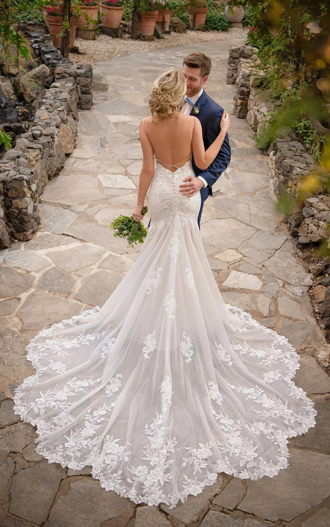 A bride in a white lace mermaid-style wedding gown with a long train stands on a stone path outdoors, facing a groom in a navy suit. The scene is set in a garden with stone walls and potted plants.